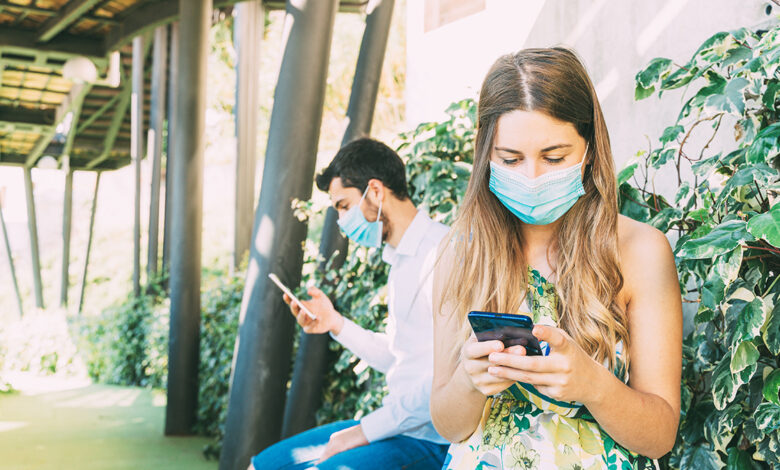 A young woman and man with face masks on sit on a bench outside with physical distance between them, while using their mobile devices - representing customer experience management in a new reality.