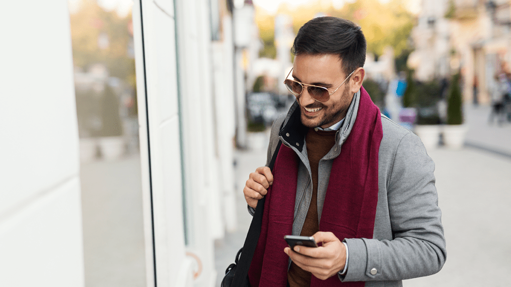 A man walks down a street holding his mobile device while looking into a store window.