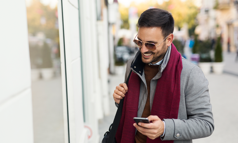 A man walks down a street holding his mobile device while looking into a store window.