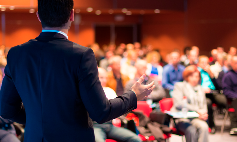 male presenter speaks to business audience, view is from behind the speaker
