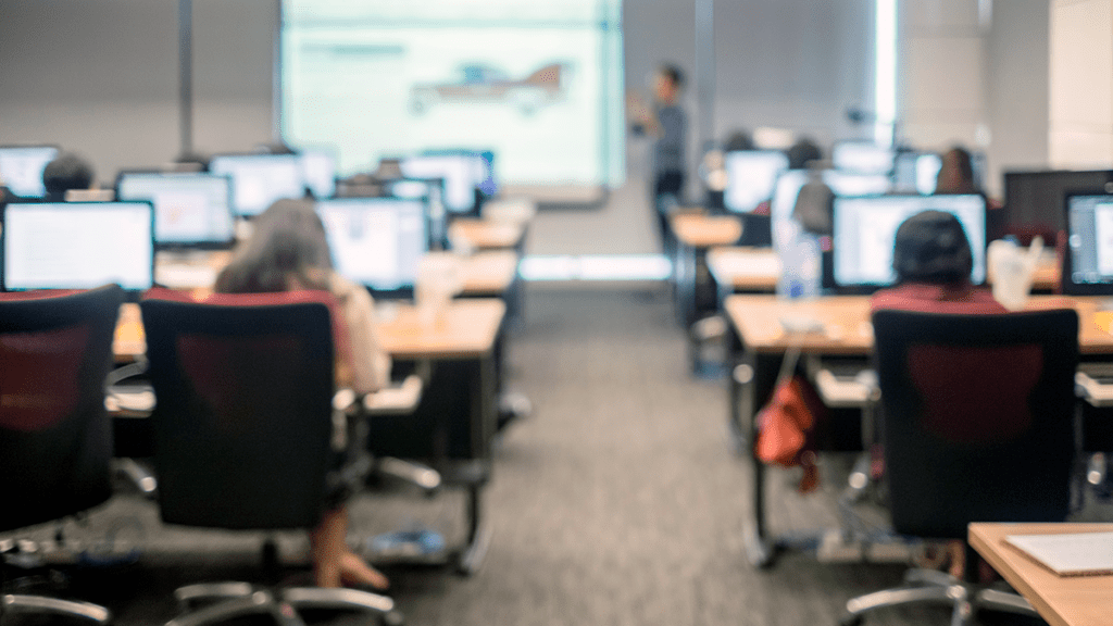 presentation room full of tables with desktop computers and chairs lined up, room is comfortably full of business people, the presenter is at the front of the room, just out of focus, and he is pointing to a large screen