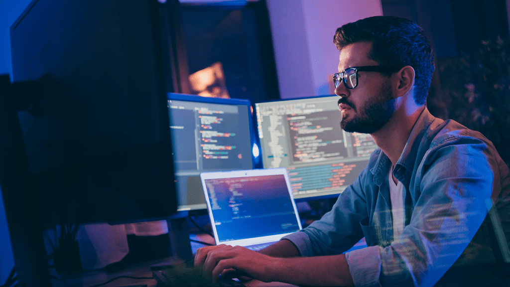 man in glasses sitting at desk looking at 4 monitors on his desk, typing on keyboard; background is dark, lighting is from monitors