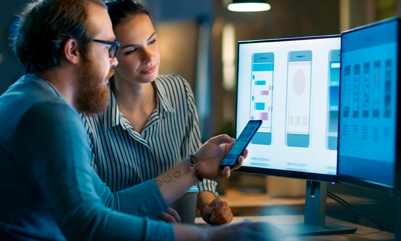 A man and a woman looking at two monitors, and the woman is comparing it to the mobile version on her phone.