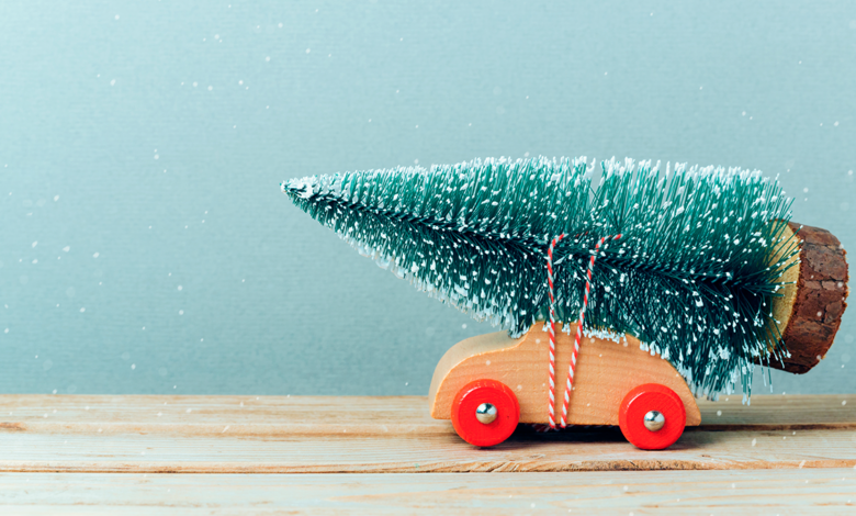 Little toy wooden car with toy Christmas tree tied to its roof appears to be traveling from right side of image to left on a wooden table top.