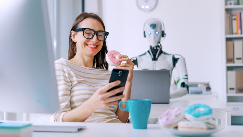 woman sitting at desk in front of laptop, but she's smiling at her mobile and eating a pink doughnut, while a robot is working at a laptop in the background