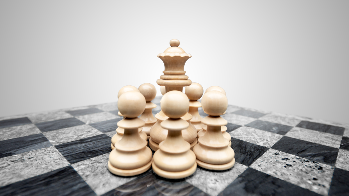 A white queen chess piece is surrounded by several white pawns, indicating protection, on a chess board.