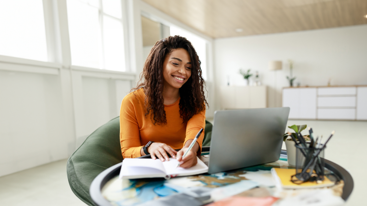 happy woman sitting on comfortable chair with laptop making notes as well; background is modern office setting