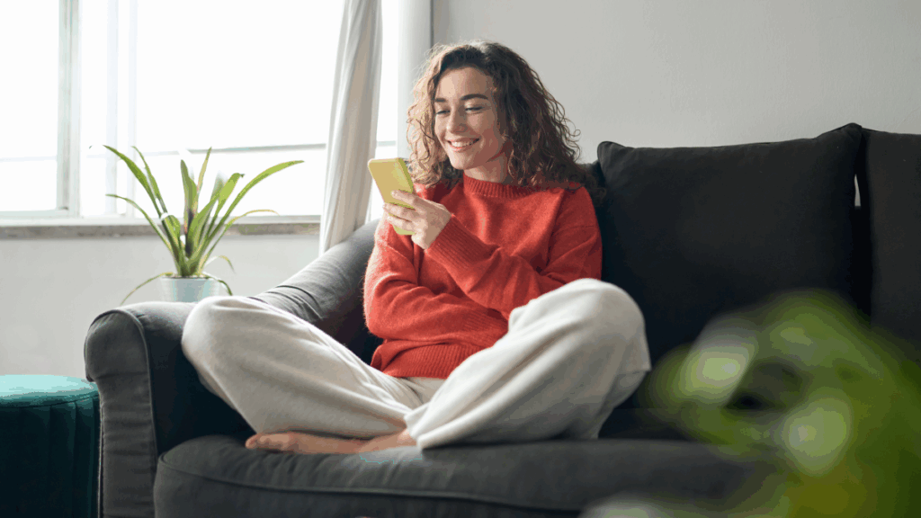 a woman sitting cross-legged on a couch, smiling, wearing an orange sweater and beige sweatpants